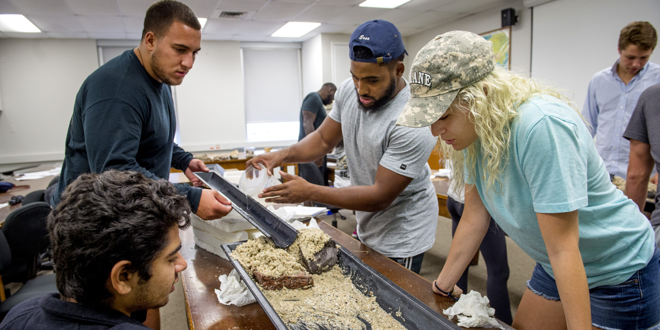 Four students stand alongside a long, narrow metal water table inside a classroom, heads down watching water and sand interact.