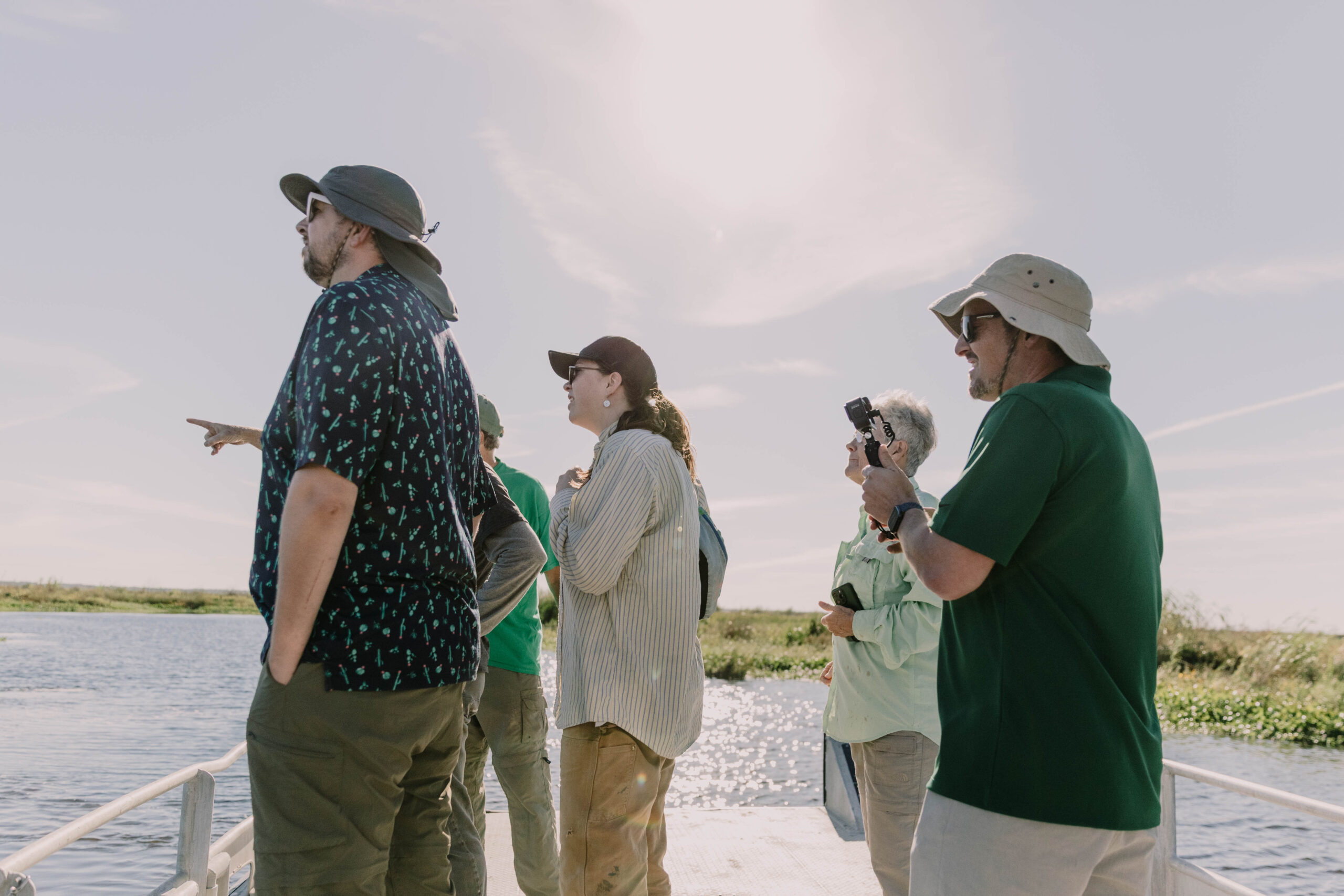 Four students stand alongside a long, narrow metal water table inside a classroom, heads down watching water and sand interact.